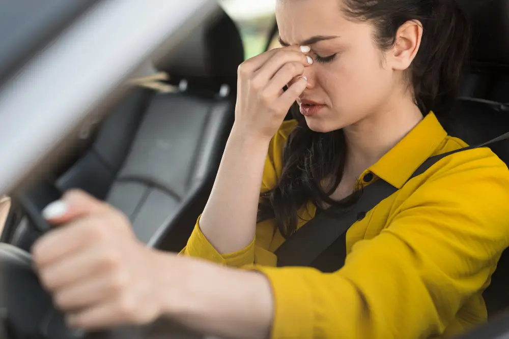 A woman in a yellow shirt pinching the bridge of her nose, showing discomfort while driving, illustrating the potential health risks from toxic chemicals in car interiors.